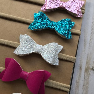May include: A collection of baby headbands featuring various bow designs. The bows come in a range of colors, including pink, turquoise, white, and magenta. Some bows have a glittery finish, while others have a smooth, leather-like texture. The headbands are attached to a neutral-colored backing.