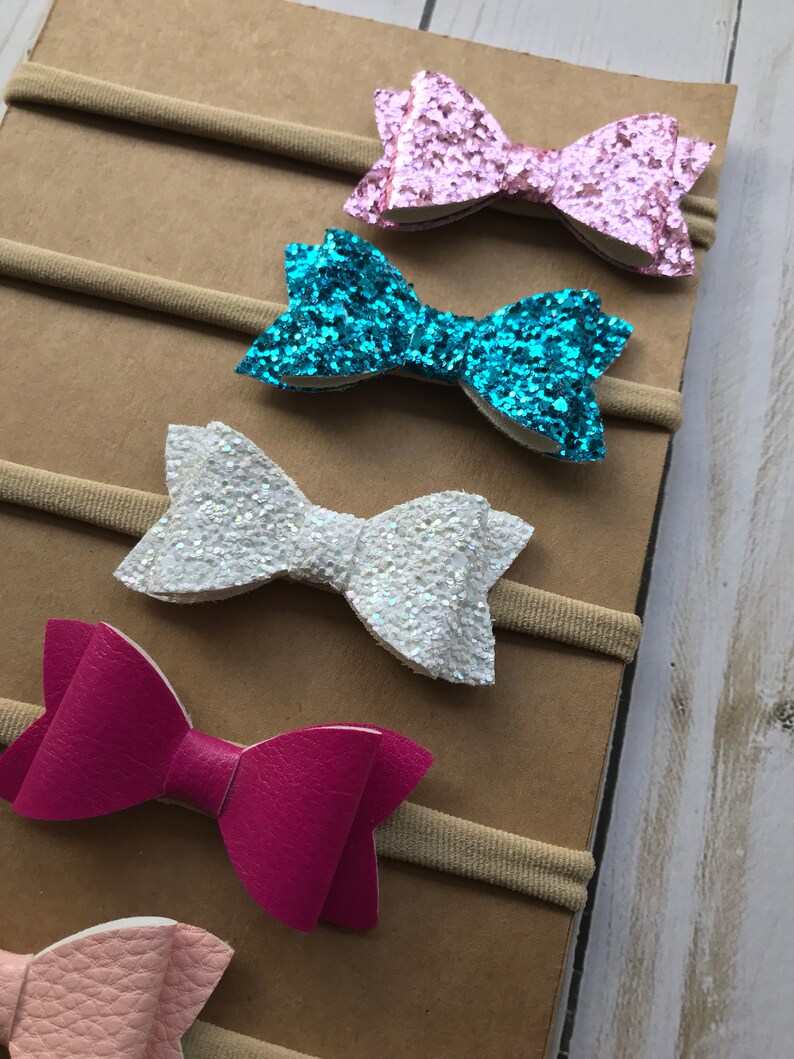 May include: A collection of baby headbands featuring various bow designs. The bows come in a range of colors, including pink, turquoise, white, and magenta. Some bows have a glittery finish, while others have a smooth, leather-like texture. The headbands are attached to a neutral-colored backing.