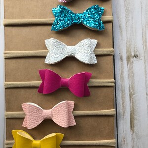May include: A collection of six baby headbands with bow accents. The bows are in various colors, including pink glitter, turquoise glitter, white, fuchsia, light pink, and yellow. Each bow is attached to a neutral-toned elastic band.