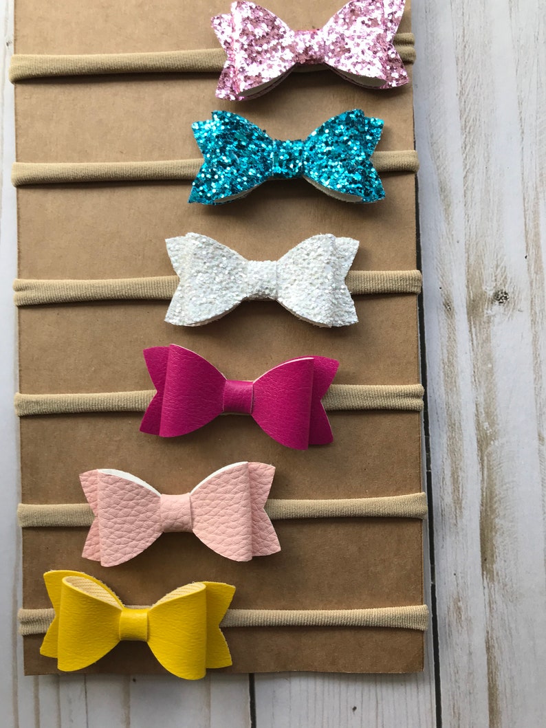 May include: A collection of six baby headbands with bow accents. The bows are in various colors, including pink glitter, turquoise glitter, white, fuchsia, light pink, and yellow. Each bow is attached to a neutral-toned elastic band.