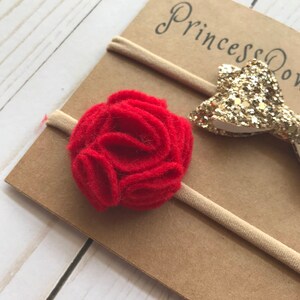 May include: A baby headband set featuring a red felt flower and a gold glitter bow. The headbands are attached to a tan elastic band and displayed on a brown card with the text "Princess Bows".