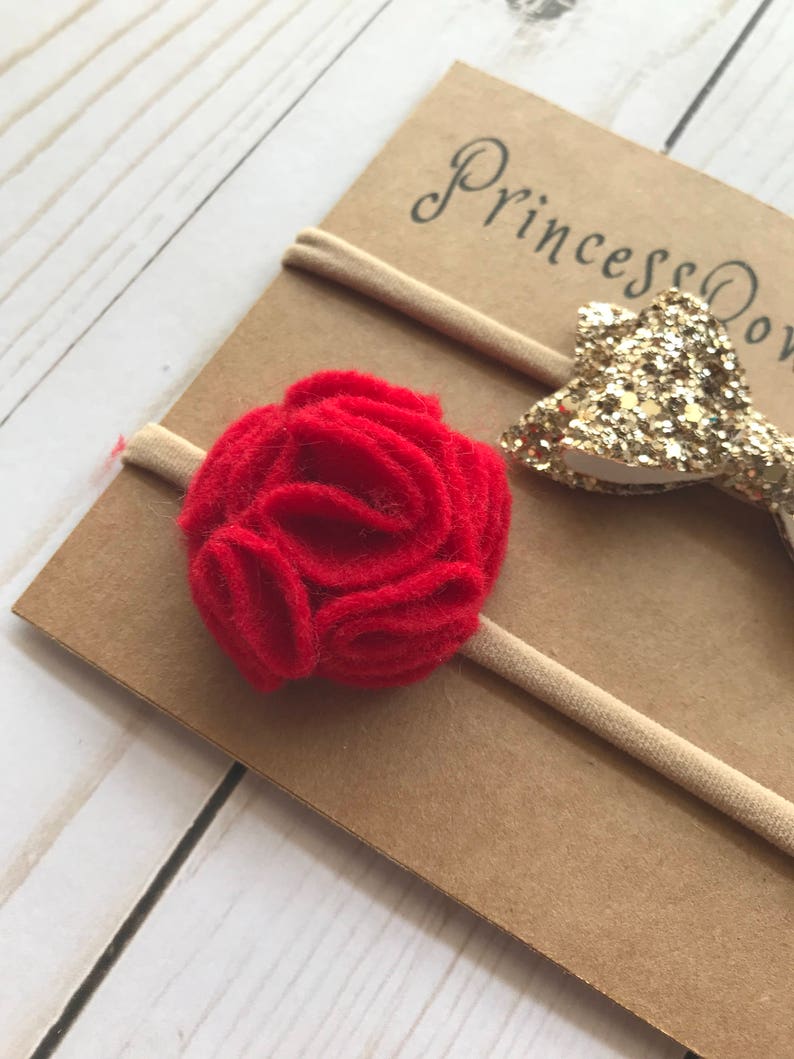 May include: A baby headband set featuring a red felt flower and a gold glitter bow. The headbands are attached to a tan elastic band and displayed on a brown card with the text "Princess Bows".