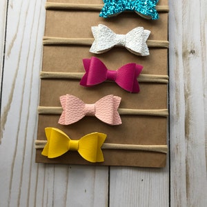 May include: A display of six colorful hair bows on elastic bands. The bows are in various colors, including pink glitter, turquoise glitter, white, fuchsia, light pink, and yellow. The bows are attached to a brown cardboard backing.