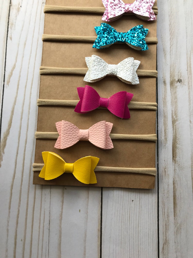 May include: A display of six colorful hair bows on elastic bands. The bows are in various colors, including pink glitter, turquoise glitter, white, fuchsia, light pink, and yellow. The bows are attached to a brown cardboard backing.