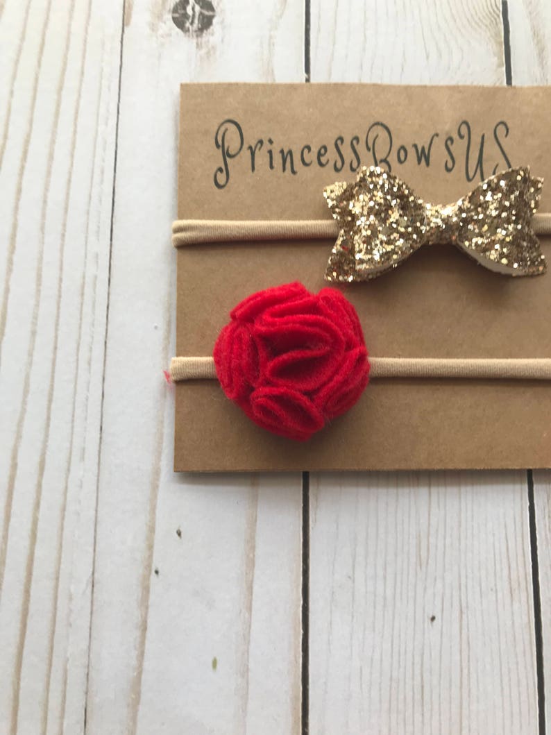 May include: Two baby headbands on a cardboard backing. One headband features a gold glitter bow, and the other has a red felt flower. The cardboard is printed with the text "Princess Bows US". The background is a light wood.