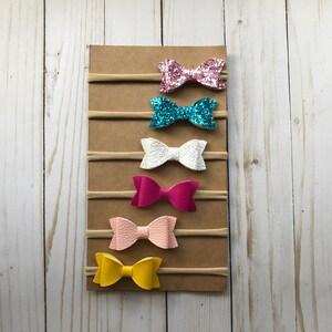 May include: A display of six colorful hair bows on elastic bands, attached to a brown cardboard backing. The bows are in various colors, including pink glitter, turquoise glitter, white, fuchsia, light pink, and yellow.