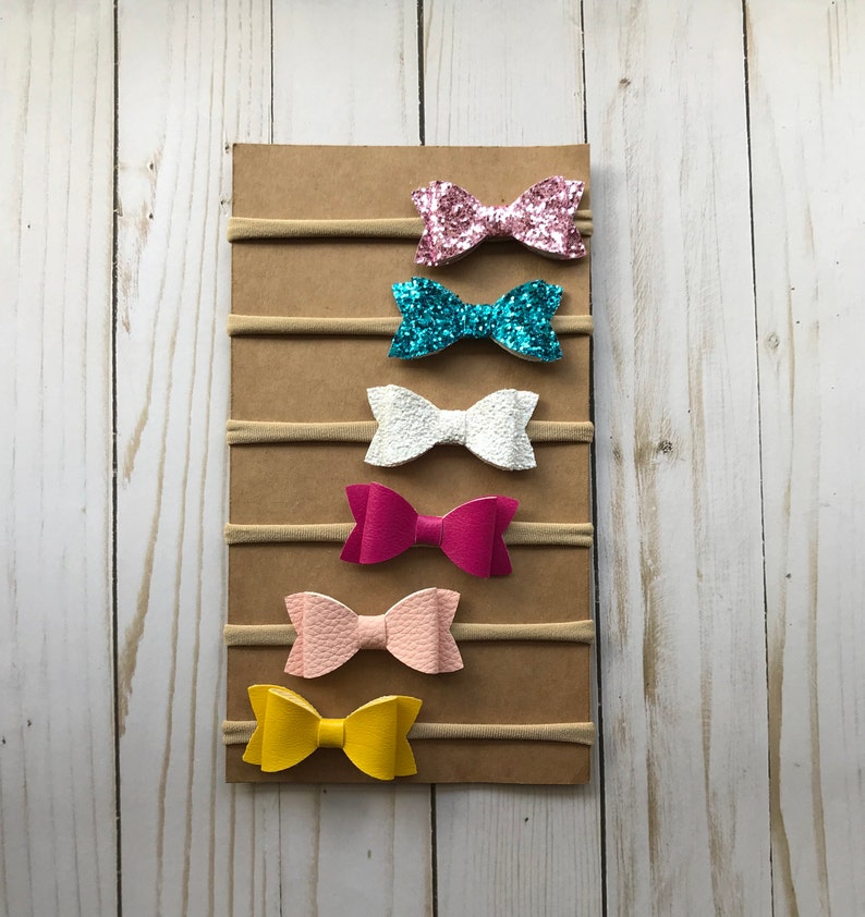 May include: A display of six colorful hair bows on elastic bands, attached to a brown cardboard backing. The bows are in various colors, including pink glitter, turquoise glitter, white, fuchsia, light pink, and yellow.