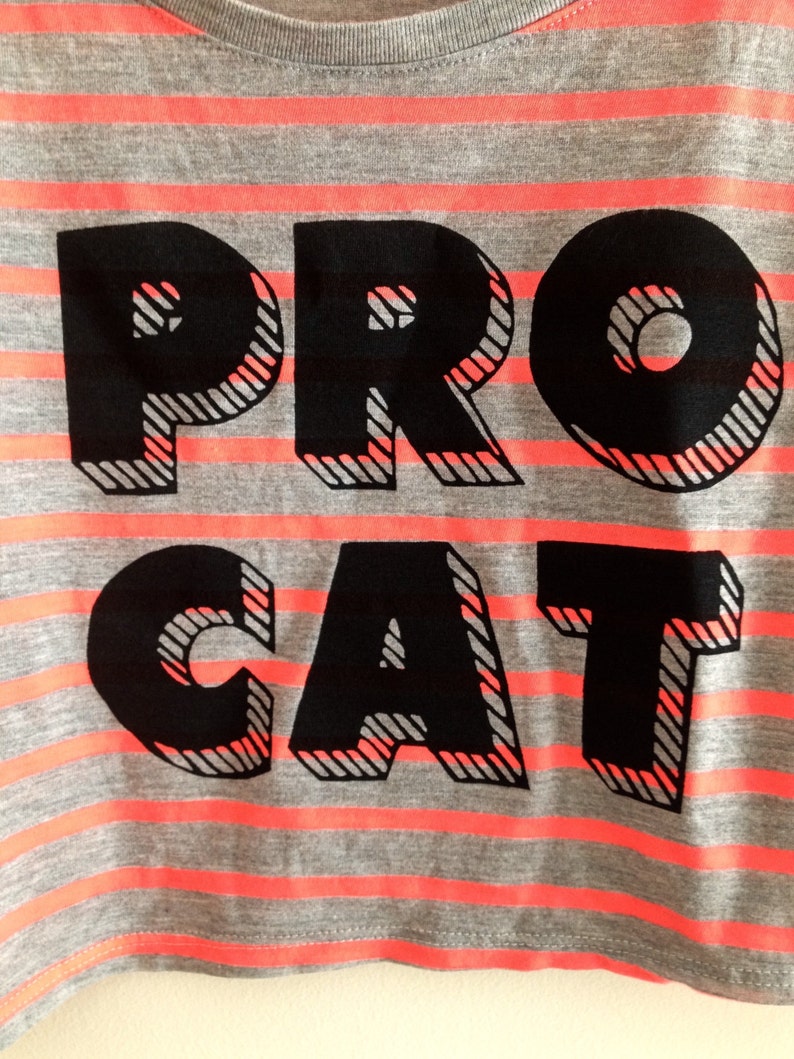 Op de afbeelding: Gekropt shirt met grijze en roze strepen en zwarte tekst die "PRO CAT" luidt.