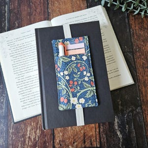 Puede incluir: Un bolsillo de libro de tela azul con un estampado floral, que contiene un bolígrafo y notas adhesivas, sujeto a un libro negro con un marcapáginas de cinta blanca. Libros abiertos en el fondo.