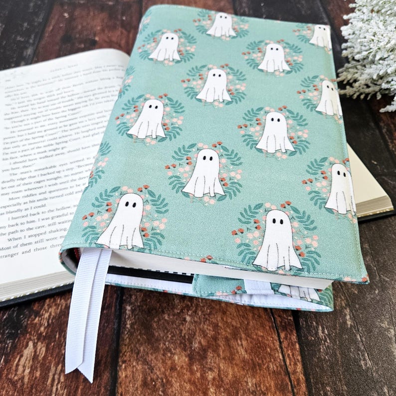 Peut inclure: Une housse de livre turquoise avec un motif de fant&ocirc;mes blancs. Les fant&ocirc;mes sont entour&eacute;s de couronnes de feuilles et de petites fleurs roses. La housse a un marque-page blanc.