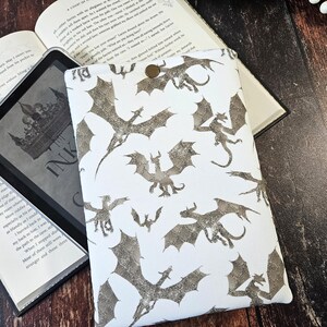 K&ouml;nnte beinhalten: Wei&szlig;e E-Reader-H&uuml;lle mit einem sich wiederholenden Muster aus silbernen Glitzerdrachen. Die H&uuml;lle hat einen Knopfverschluss.