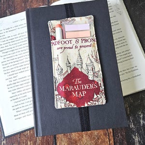 May include: A fabric pocket with the text "PADFOOT & PRONG are proud to present The MARAUDER'S MAP" is attached to a black notebook. The pocket contains a pink pen and two sticky notes. The design features a castle illustration.