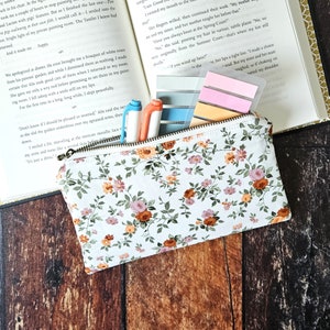 May include: A white zippered pouch with a floral pattern featuring small pink, orange, and yellow flowers. The pouch is lying open on a wooden surface with three pens and several colorful sticky notes inside.