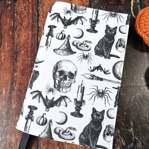 May include: A white fabric book cover with a black and white Halloween themed pattern. The pattern includes bats, skulls, spiders, snakes, cats, moons, candles, and witches' hats.