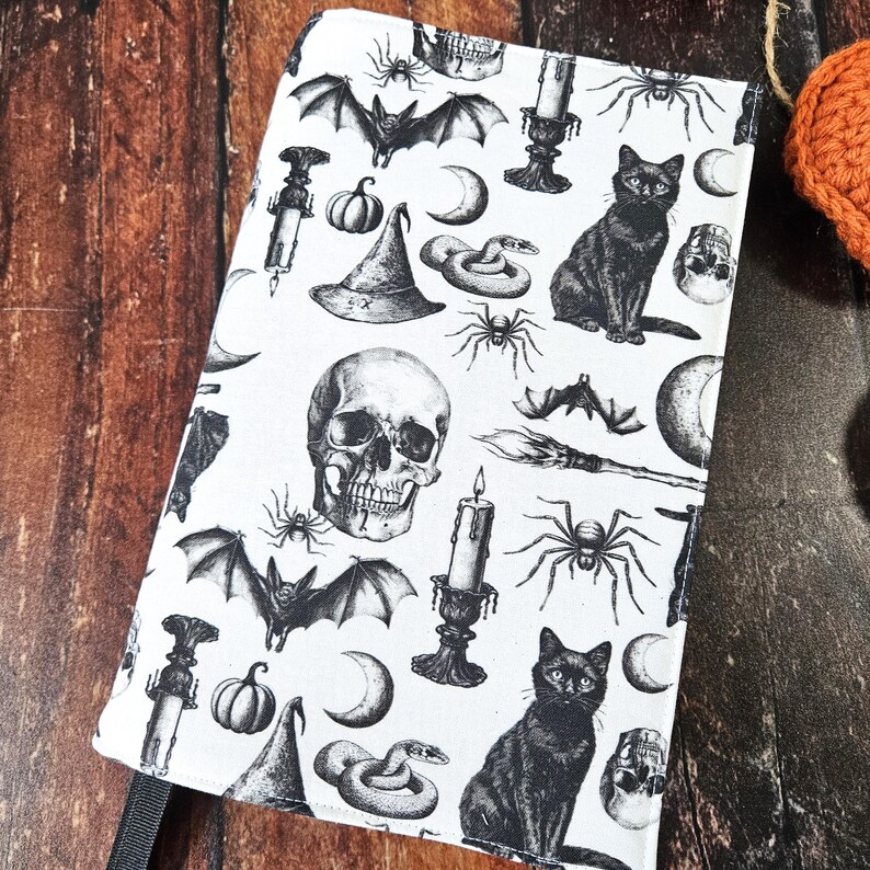 May include: A white fabric book cover with a black and white Halloween themed pattern. The pattern includes bats, skulls, spiders, snakes, cats, moons, candles, and witches' hats.