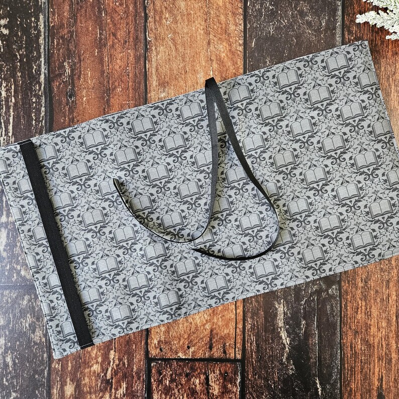 May include: A gray fabric book roll with a black ribbon tie and a repeating pattern of open books. The fabric has a subtle texture.