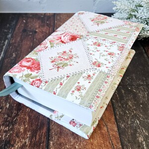 K&ouml;nnte beinhalten: Ein floral gemusterter Buchumschlag mit Patchwork-Design. Der Umschlag zeigt eine Mischung aus wei&szlig;en, rosa und gr&uuml;nen Blumenmustern. Ein hellgr&uuml;nes Lesezeichenband ist sichtbar. Das Buch liegt auf einer dunklen Holzoberfl&auml;che.