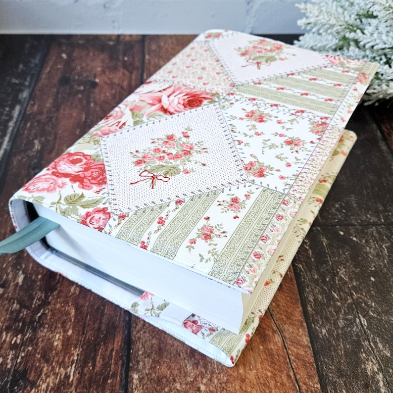 K&ouml;nnte beinhalten: Ein floral gemusterter Buchumschlag mit Patchwork-Design. Der Umschlag zeigt eine Mischung aus wei&szlig;en, rosa und gr&uuml;nen Blumenmustern. Ein hellgr&uuml;nes Lesezeichenband ist sichtbar. Das Buch liegt auf einer dunklen Holzoberfl&auml;che.