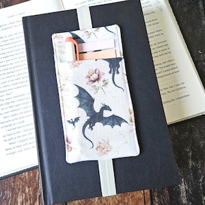 Puede incluir: Marcapáginas de tela con bolsillo, con un diseño de dragón y flores. El marcapáginas contiene un bolígrafo y notas adhesivas, y está colocado en un libro. El diseño incluye un dragón, flores y un fondo crema.