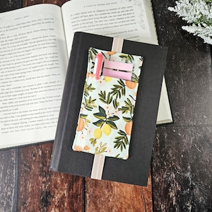 May include: A hardback book with a black cover and a floral bookmark. The bookmark has a pattern of lemons, leaves, and flowers in yellow, green, and orange. A pink pen and sticky notes are in the pocket.