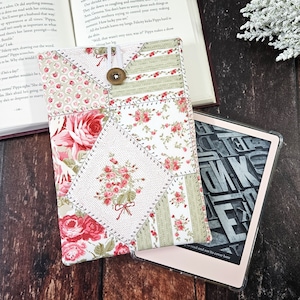 May include: A white and green patchwork fabric e-reader cover with pink roses and a button closure. The cover is lying on a wooden surface next to a black and white e-reader.