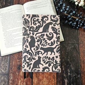 May include: A pink book cover with a black and white damask pattern featuring black cats. The cover has a black bookmark attached.