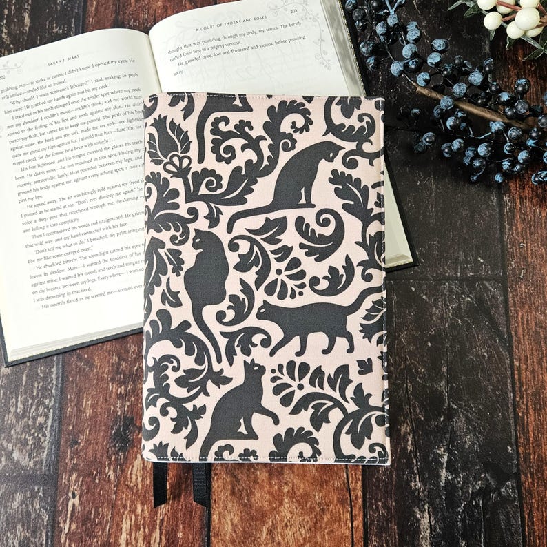 May include: A pink book cover with a black and white damask pattern featuring black cats. The cover has a black bookmark attached.