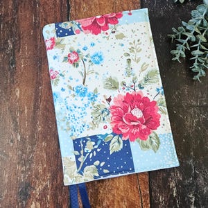 May include: A floral book cover with a patchwork design. The cover features large pink and red flowers, blue accents, and a light blue border. A navy blue ribbon bookmark is attached.