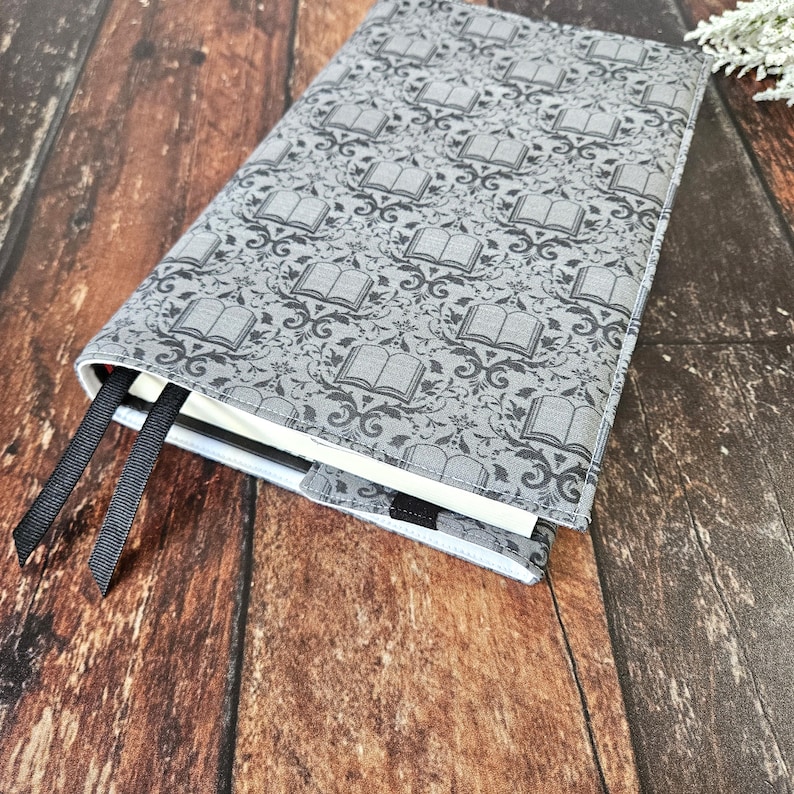 May include: A gray book cover with a repeating pattern of black books and floral designs. The cover has black ribbon bookmarks attached.