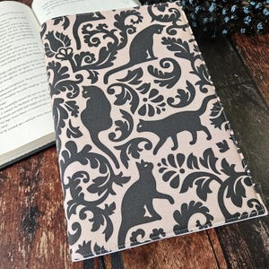 May include: A book cover with a black and white damask pattern featuring black cats. The background is a light beige colour.
