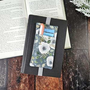 May include: A grey fabric bookmark with white floral print and a pocket for a pen and sticky notes. The bookmark is attached to a black book.