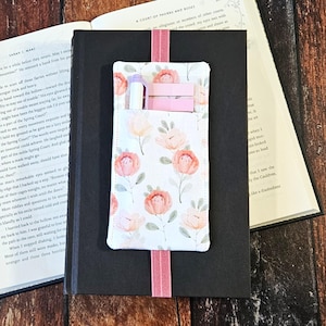 May include: A fabric bookmark with a pocket, holding a pen and pink sticky notes, rests on a closed black book. The bookmark features a white background with pink and peach flower designs and a pink elastic band.