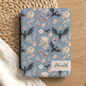 Puede incluir: Una funda azul para lector de libros electrónicos con un estampado floral y de dragones. El diseño presenta dragones negros, flores beige y acentos rosas. Un rectángulo blanco con el nombre "Meredith" en escritura cursiva está en la funda.