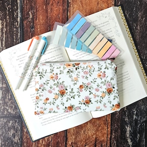 May include: A white zippered pouch with a floral pattern featuring small pink and orange flowers. The pouch is lying open on a book with colorful sticky notes.