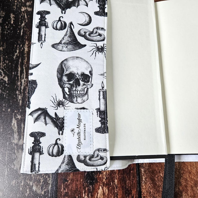 May include: A white notebook cover with a black and white Halloween-themed print. The print features skulls, bats, snakes, candles, and a witch's hat. The cover has a label that reads "Elizabeth Mayfair Handmade".