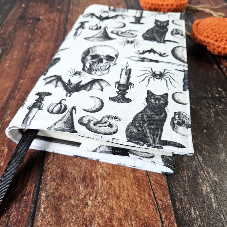 May include: A white book cover featuring a black and white print of Halloween themed images, such as bats, cats, skulls, snakes, pumpkins, and candles. The cover has a black ribbon bookmark.