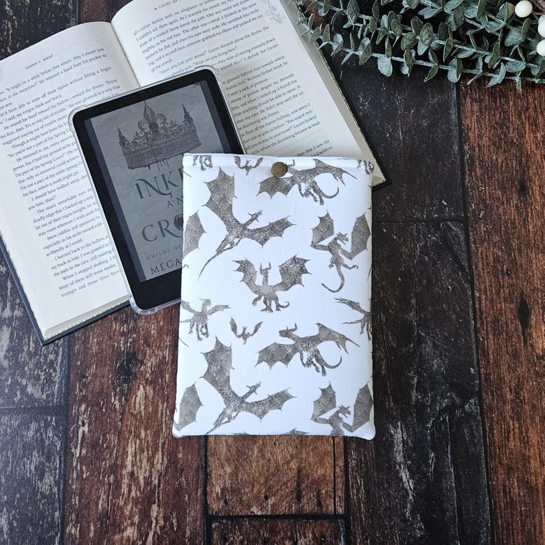 K&ouml;nnte beinhalten: Wei&szlig;e E-Book-H&uuml;lle mit einem sich wiederholenden Muster aus grauen Drachen-Silhouetten. Die H&uuml;lle hat einen goldenen Knopfverschluss. Der Text "Ink and a Crown" ist auf dem E-Book-Reader im Hintergrund sichtbar.