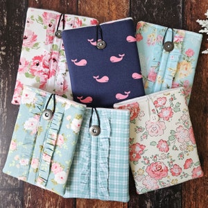 May include: Six different fabric covers for a phone or small notebook. The covers are in various floral patterns, including pink and white roses, blue and white flowers, and a light blue and white plaid. One cover features a navy blue background with white whales and pink whale silhouettes.