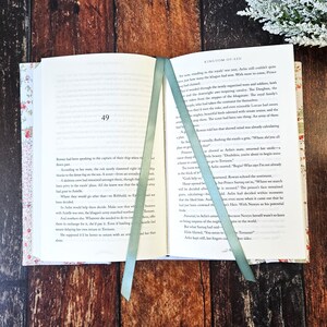 K&ouml;nnte beinhalten: Offenes Buch mit floralem Einband und zwei hellgr&uuml;nen Leseb&auml;ndchen. Das Buch ist auf Seite 49 aufgeschlagen, mit Text sichtbar. Der Titel "Kingdom of Ash" ist oben auf der Seite zu sehen.
