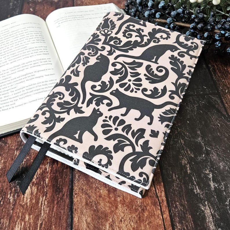 May include: A book cover with a black and white damask pattern featuring black cats. The cover is made of a soft, beige fabric and has a white border.