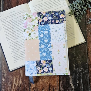 May include: A rectangular fabric book cover featuring a patchwork design of various floral patterns in shades of navy blue, light blue, soft pink, and white. The cover includes a small blue ribbon bookmark attached to the bottom edge of the spine.