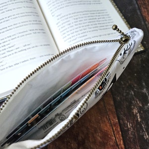 K&ouml;nnte beinhalten: Eine wei&szlig;e Rei&szlig;verschlusstasche mit einem schwarz-wei&szlig;en Druck eines Wesens mit gro&szlig;en Augen. Die Tasche ist offen und enth&auml;lt mehrere farbige Karteikarten.