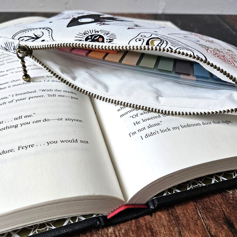 K&ouml;nnte beinhalten: Eine wei&szlig;e Canvas-Rei&szlig;verschlusstasche mit einem schwarz-wei&szlig;en grafischen Design. Die Tasche ist ge&ouml;ffnet und ein Buch ist sichtbar mit dem Text "Of what morning" und "He lowered the".