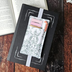 Puede incluir: Un marcapáginas de tela blanca con un patrón floral y un bolsillo para guardar notas adhesivas y un bolígrafo. El marcapáginas está unido a un libro de tapa dura negro.