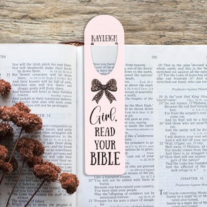 May include: A pink bookmark with the name "KAYLEIGH" at the top. Below is a leopard print bow graphic and the words "Girl, READ YOUR BIBLE" in a bold, black font. The bookmark is placed in an open book.