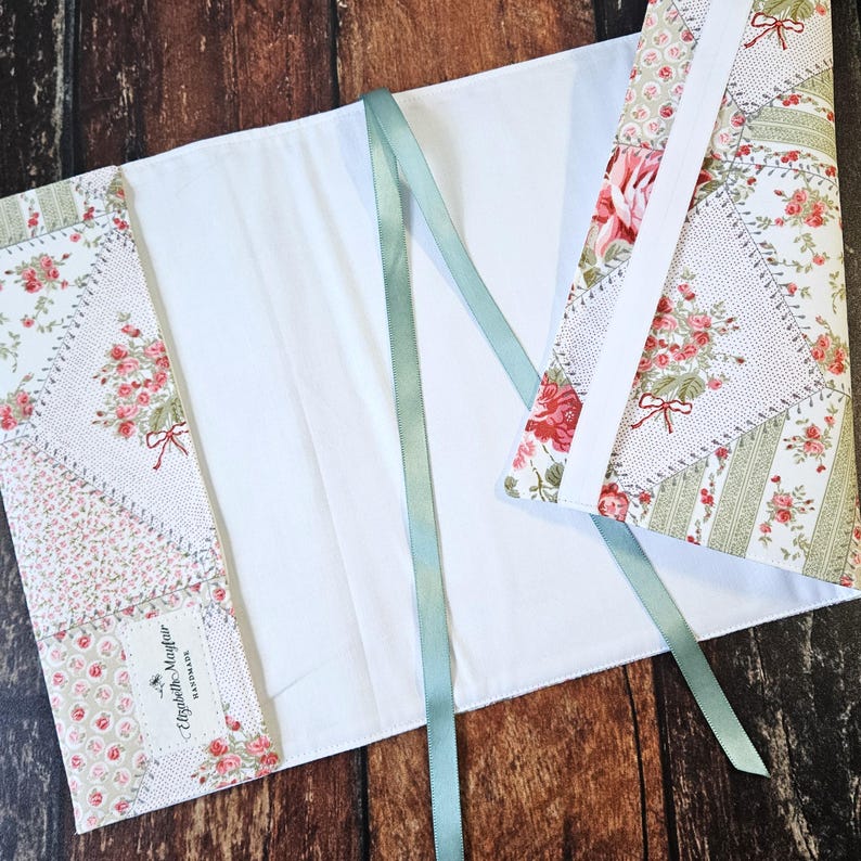 K&ouml;nnte beinhalten: Ein handgefertigter Buchumschlag mit floralem Patchwork-Design. Der Umschlag hat eine wei&szlig;e Innenseite und einen hellgr&uuml;nen Bandverschluss. Der Patchwork-Stoff zeigt rosa Rosen und gr&uuml;ne Akzente. Das Etikett lautet "Elizabeth Mayfair Handmade".