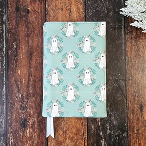 Peut inclure: Une couverture de carnet bleu sarcelle avec un motif de fant&ocirc;mes blancs. Les fant&ocirc;mes sont entour&eacute;s de couronnes de feuilles et de fleurs. La couverture a un marque-page en ruban blanc.