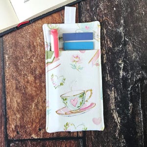 May include: A floral print book pocket with a pink pen and two blue sticky notes in the pocket. The pocket features a teacup design with a pink heart and a white loop at the top.