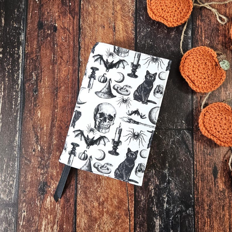 May include: A white book cover featuring a black and white illustration of a skull, bats, cats, spiders, candles, and other Halloween-themed imagery.