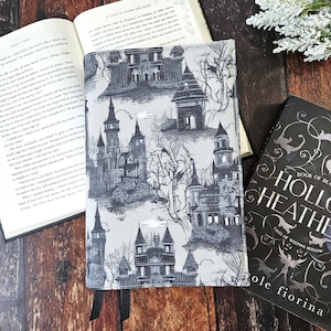 May include: A book cover with a black and white pattern of spooky houses and trees. The cover is made of fabric and has a gray background. The book is titled "Hollow Heath: Tales of Weeping Hollow" by Cole Fiorina.